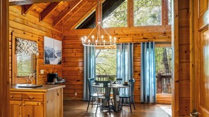 Interior de uma cabana de madeira com área de jantar, bancada de cozinha e uma janela grande com cortinas.