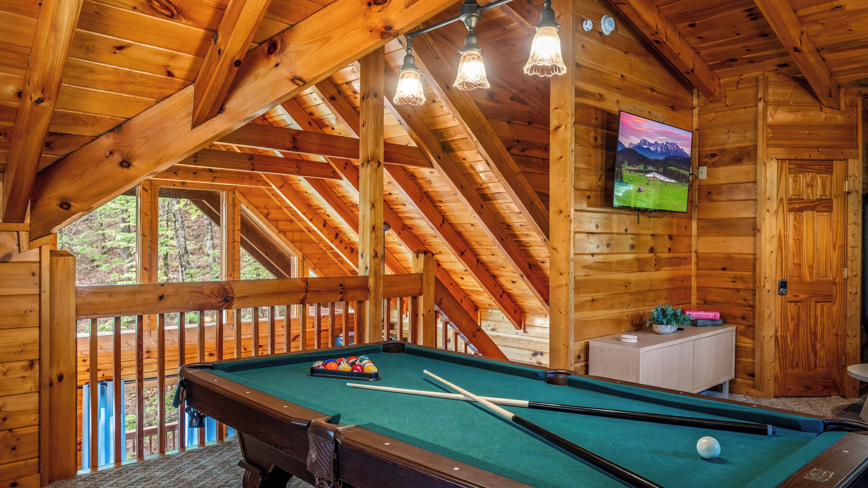 A wooden interior with a pool table, a television mounted on the wall, and a wooden deck outside.