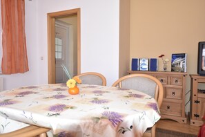 A room with a floral tablecloth, wooden chairs, a small orange vase with flowers, a wooden dresser, and a framed picture on the wall. - Schones Ferienhaus im Nationalpark Granitz (Zirkow)