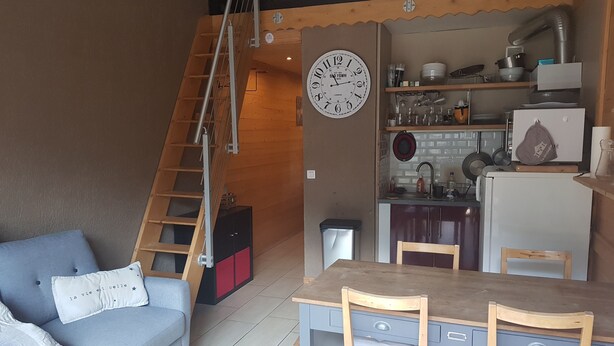Un espacio habitable compacto con sofá, una mesa de comedor con sillas, zona de cocina con fregadero y estufa, y una escalera de madera.