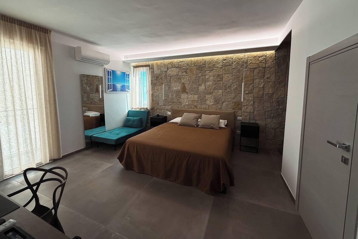 A modern bedroom with a stone accent wall, a bed with brown bedding, a blue daybed, and a desk with a chair.