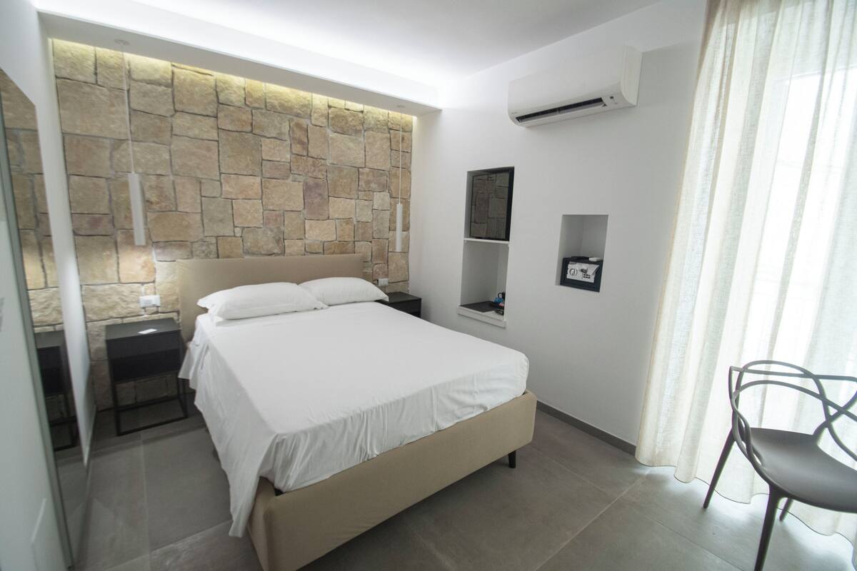 A bedroom with a stone wall, a bed, a nightstand, a chair, and a window with curtains.