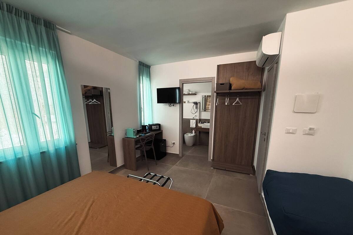 A hotel room with a bed, a desk, a chair, a television, a wardrobe, and a bathroom visible through an open door.
