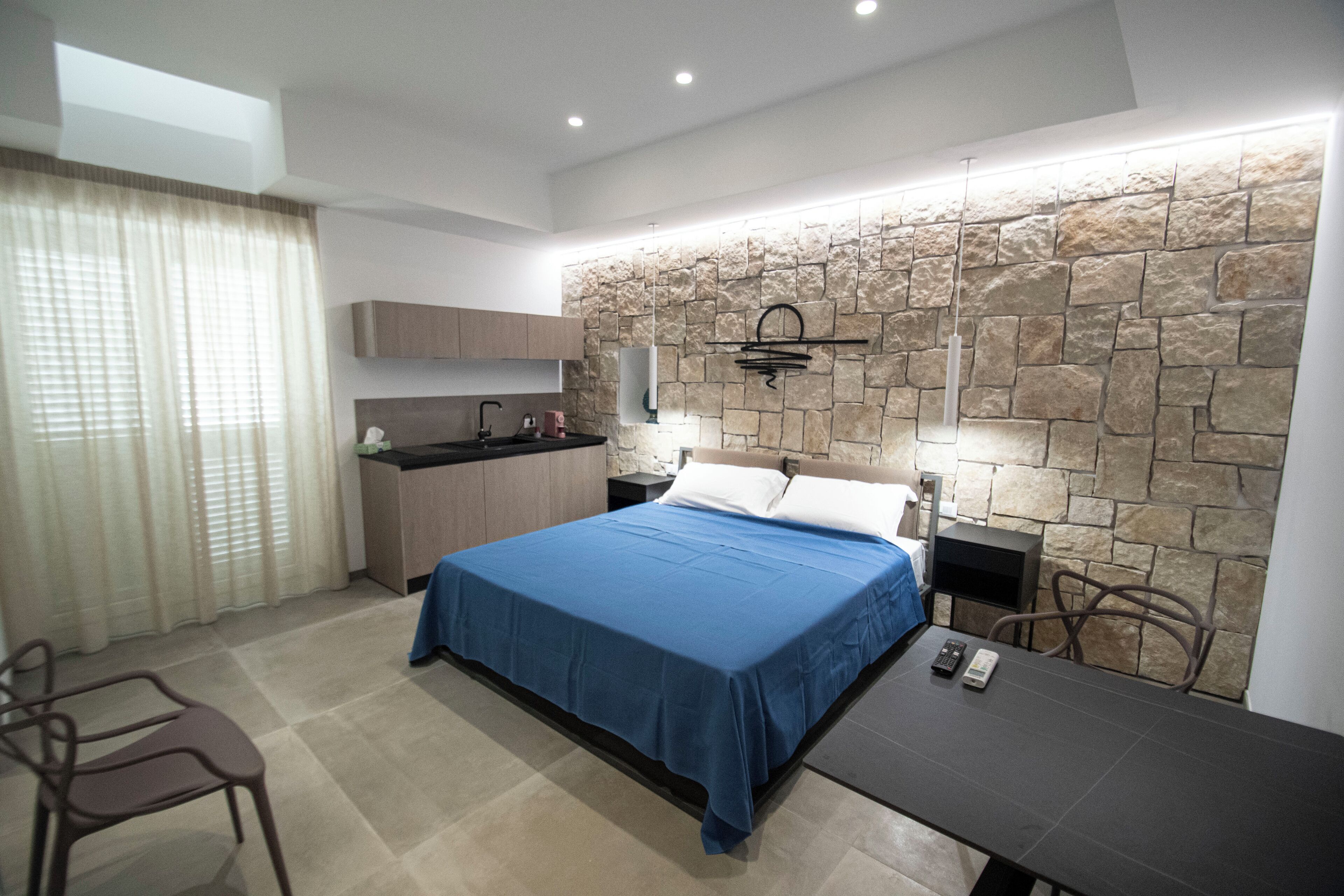 A modern bedroom with a stone accent wall, a bed with blue bedding, a small kitchenette, and a dining area with a table and chairs.