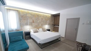 A bedroom with a bed, a blue sofa, a stone wall, a wooden wardrobe, and a door. - my Vesta Suites (Vieste)