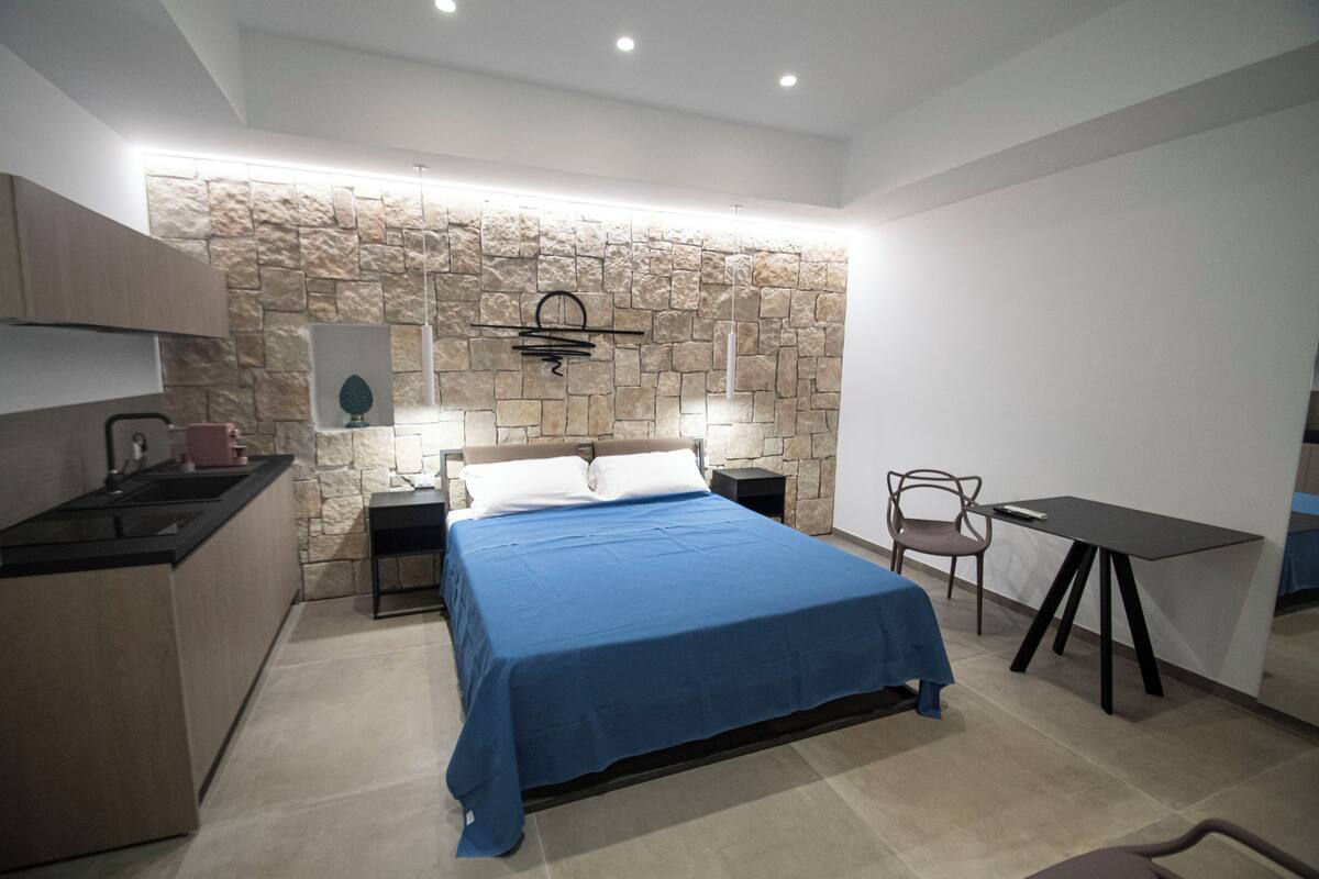 A bedroom with a bed, a desk, a chair, a nightstand, and a stone wall.