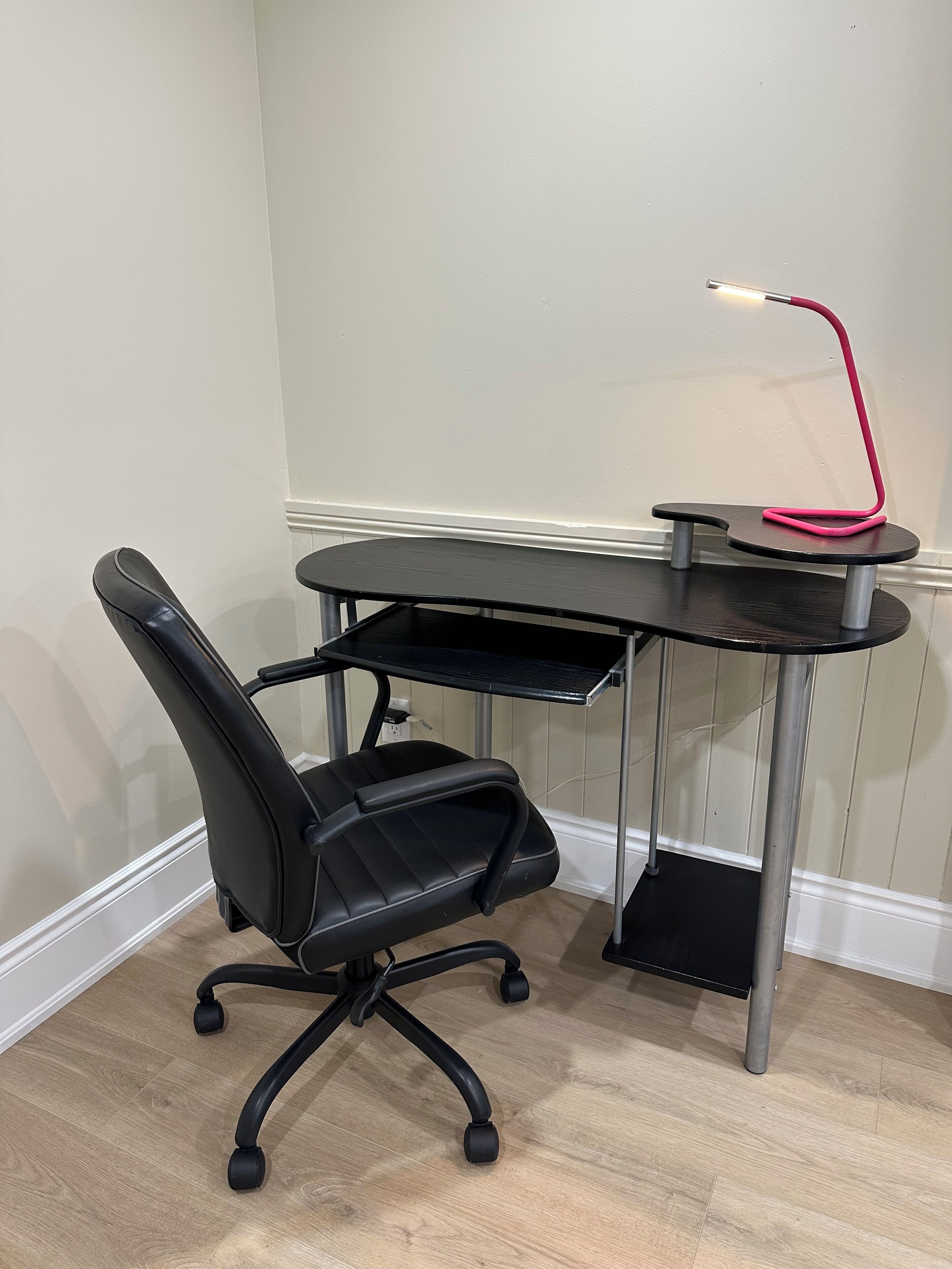Ein schwarzer Bürostuhl mit fünf Rollen, ein schwarzer Schreibtisch mit abgerundeter Kante und eine rote Lampe mit Metallfuß.