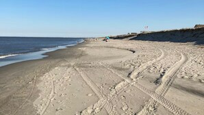Une plage de sable avec des empreintes de pas et des traces de pneus, un ciel dégagé et un drapeau flottant sur une structure visible au loin.