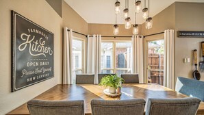 A dining area with a wooden table, four grey upholstered chairs, a potted plant, and a sign on the wall reading "FARMHOUSE Kitchen OPEN DAILY Please Seat Yourself NOW SERVING."
