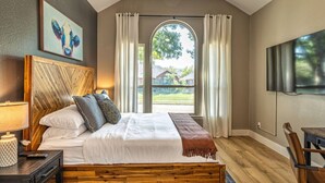 A bedroom with a large bed, a wooden headboard, a TV, and a view of a house through a window.