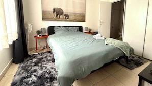 A bedroom with a large bed, a bedside table with a lamp, a wall-mounted fan, and a painting of elephants.