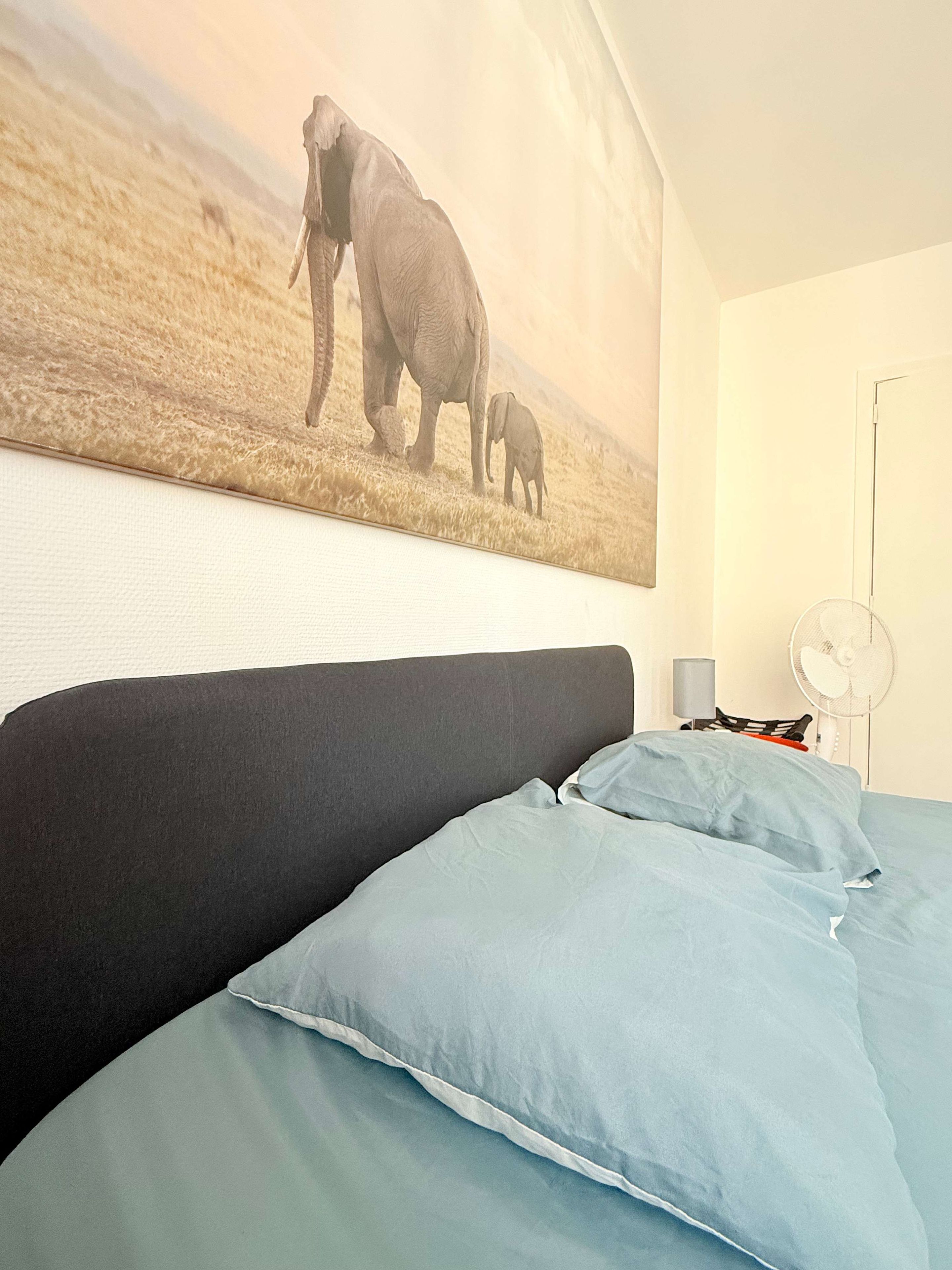 Une chambre à coucher avec un lit, des oreillers, un ventilateur fixé au mur et un grand tableau représentant des éléphants.