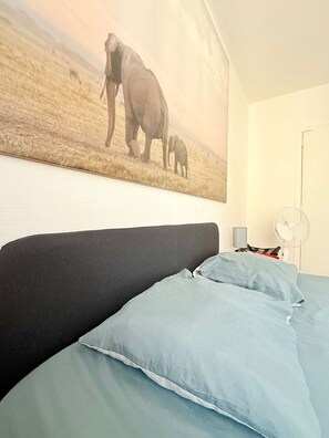 Un dormitorio con cama, almohadas, un ventilador de pared y un cuadro grande de elefantes.