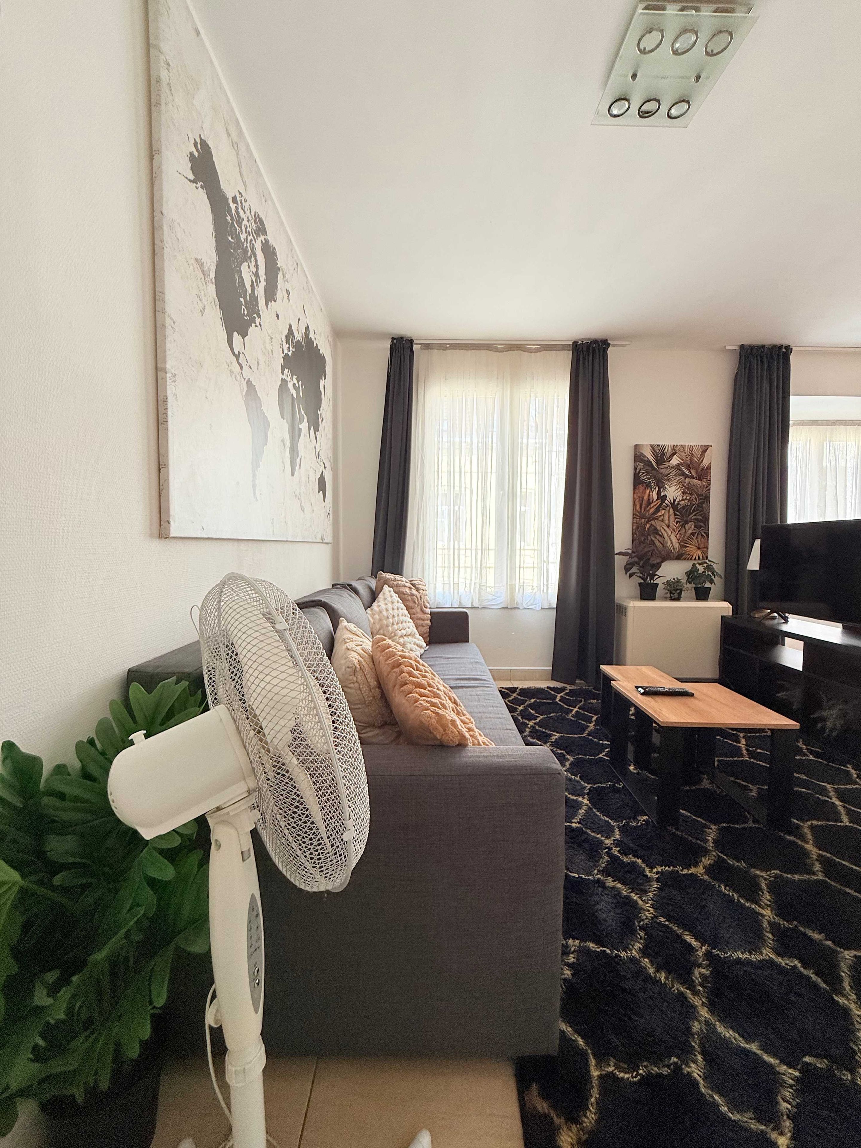 Een woonkamer met een grijze bank, een klein houten tafeltje, een witte ventilator en een groot abstract schilderij aan de muur.