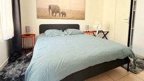 Una camera da letto con un letto, una ventola a parete, un piccolo tavolino con una lampada, un quadro raffigurante degli elefanti e una porta.