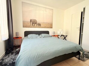 A bedroom with a bed, a wall-mounted fan, a small table with a lamp, a painting of elephants, and a door. - Bright & Refined Apartment in the Heart of the EU Quarter (Brussels)
