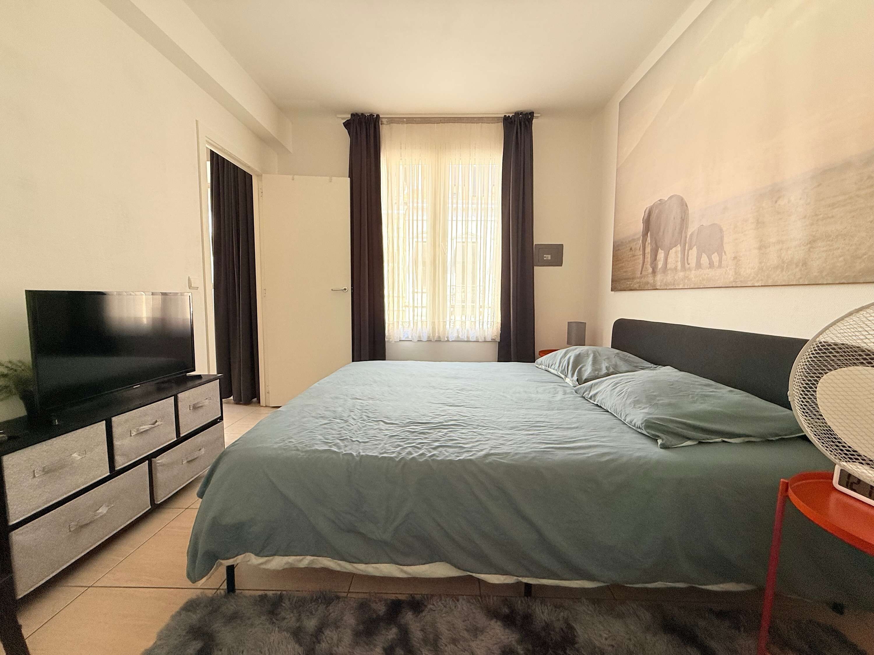 Een slaapkamer met een bed, een tv op een dressoir, een schilderij aan de muur en een raam met gordijnen.