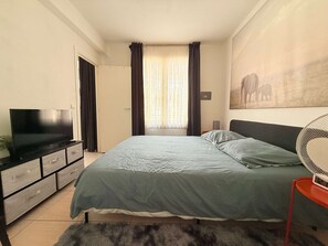 Ein Schlafzimmer mit einem Bett, einem Fernseher auf einer Kommode, einem Gemälde an der Wand und einem Fenster mit Vorhängen.