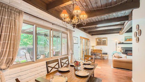 Uma área de jantar com mesa e cadeiras de madeira, um candelabro e vista para o exterior através de grandes janelas.