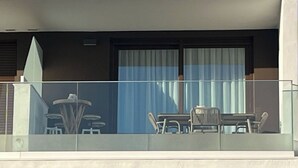 Un edificio de varios pisos con un balcón en el segundo piso. El balcón tiene una mesa, sillas, una sombrilla pequeña y un ventanal con cortinas.