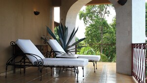 A covered patio with two white cushioned lounge chairs, a large agave plant, and a view of trees through an archway.