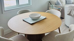 A dining area with a round wooden table, four chairs, and a white bowl on a napkin. A window offers a view of a beach and buildings.