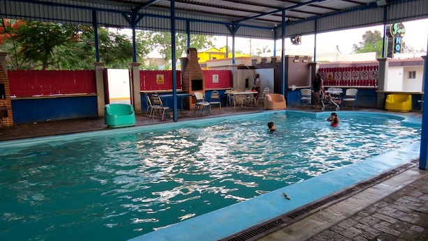 En inomhuspool med ett tak som delvis är övertäckt, omgiven av stolar och bord, och en synlig byggnad med en skylt i bakgrunden.