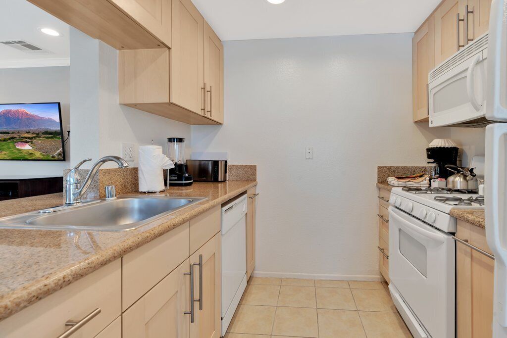 A compact kitchen with a granite countertop, stainless steel sink, white appliances, and wooden cabinets.