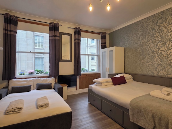 A hotel room with a bed, a sofa, a desk, and a wardrobe. There are curtains on the windows, and a mirror on the wall.