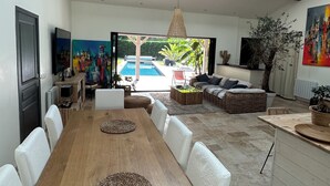 A spacious living area with a wooden dining table, white chairs, a sofa, and a large window overlooking a pool. - Indonesian villa (Lacanau)