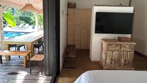 A bedroom with a large bed, a television, and a wooden bench. There is a sliding door leading to an outdoor area with a pool and lounge chairs. - Indonesian villa (Lacanau)