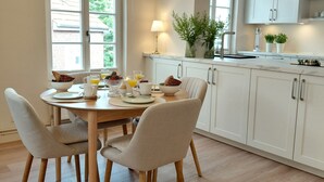 Eine moderne Küche mit weißen Schränken, einem Holzespeisetisch und Stühlen. Eine Lüster mit mehreren Leuchten, ein Fenster mit Blick ins Grüne und eine Arbeitsplatte mit einer Pflanze.