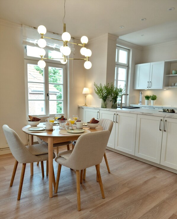 A modern kitchen with white cabinets, a wooden dining table, and chairs. There is a chandelier with multiple lights, a window with a view of greenery, and a countertop with a plant.