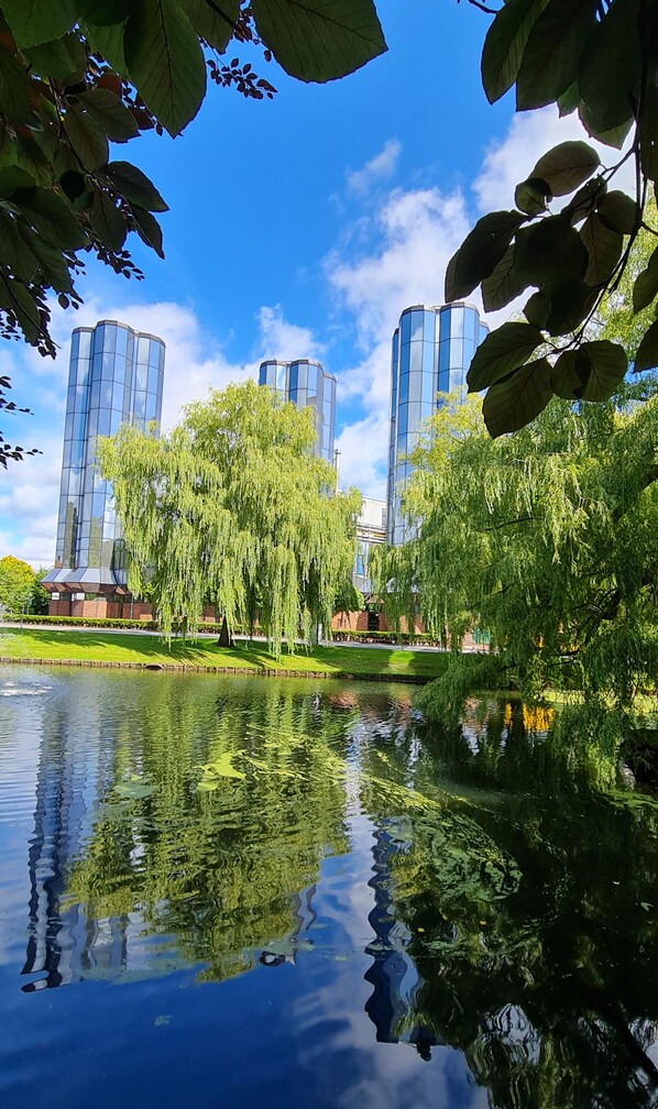 Un parc paisible avec un étang dans lequel se reflètent les bâtiments, entouré d’une végétation luxuriante et d’une architecture moderne.