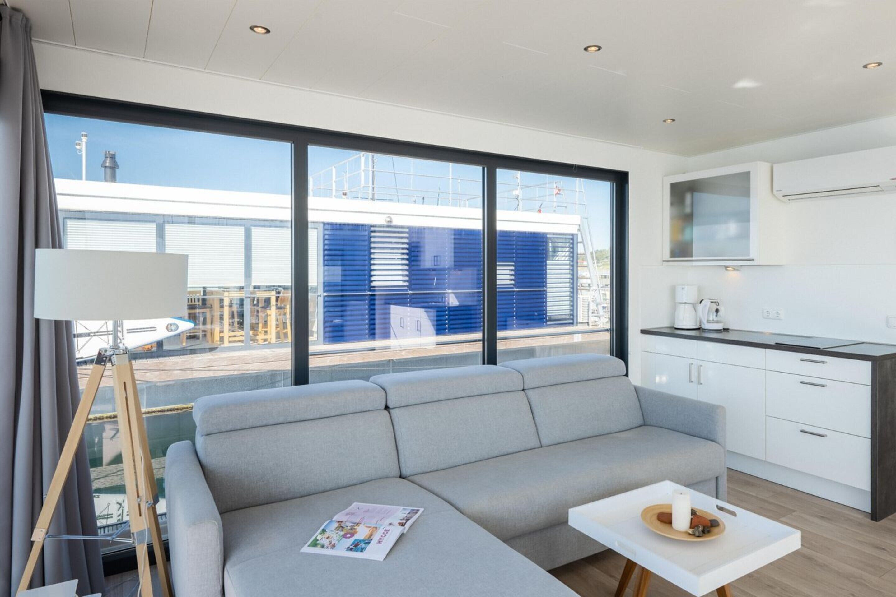 Une pièce moderne et bien éclairée, avec un canapé gris, une petite table sur laquelle se trouve un plateau, et une grande fenêtre donnant sur un immeuble aux volets bleus.