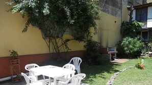 Eine Terrasse mit weißem Tisch und Stühlen, umgeben von Grünpflanzen und einer gelben Gebäudefassade.