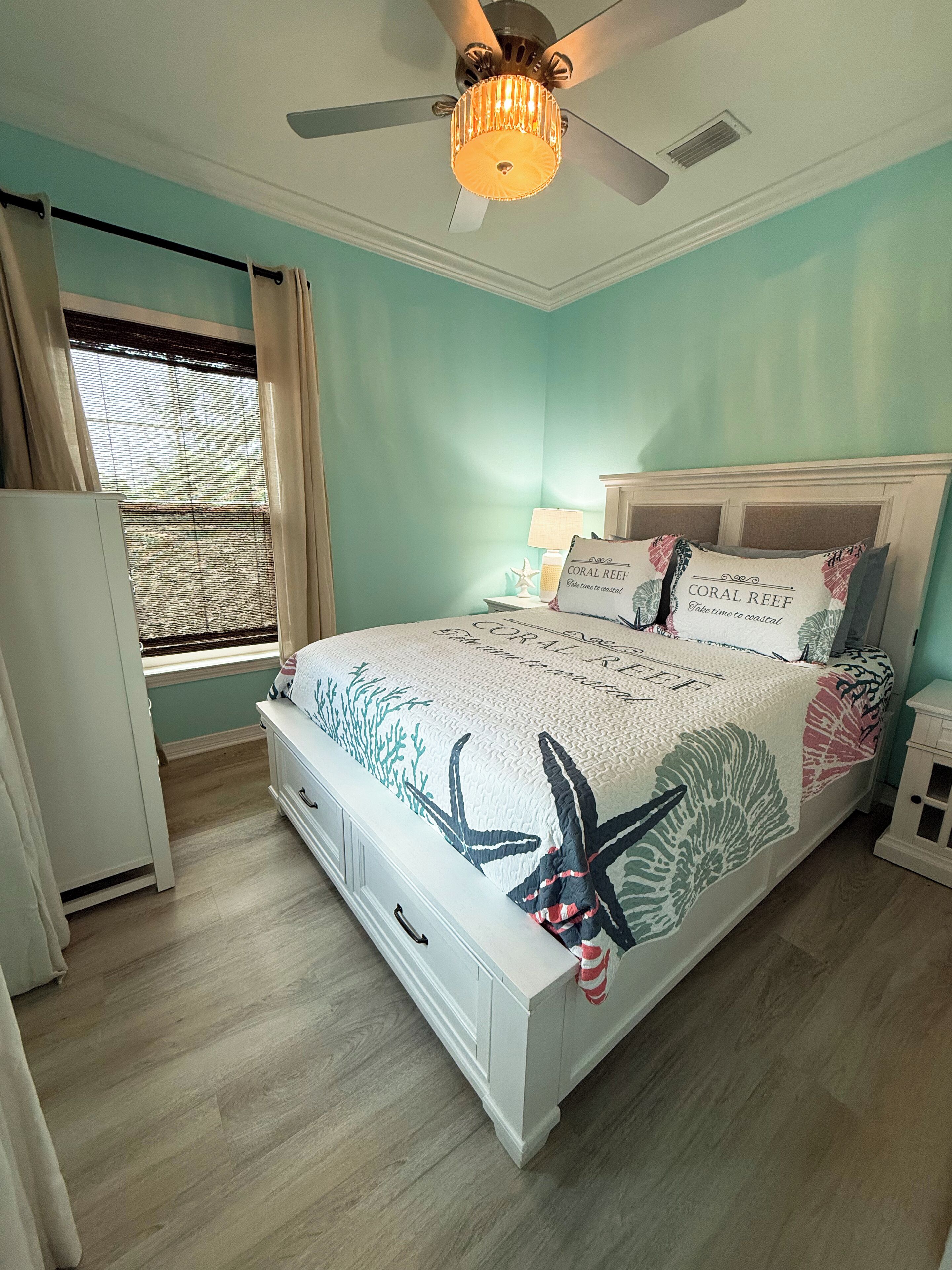 A bedroom with a bed, a ceiling fan, a window with curtains, and a nightstand with a lamp.