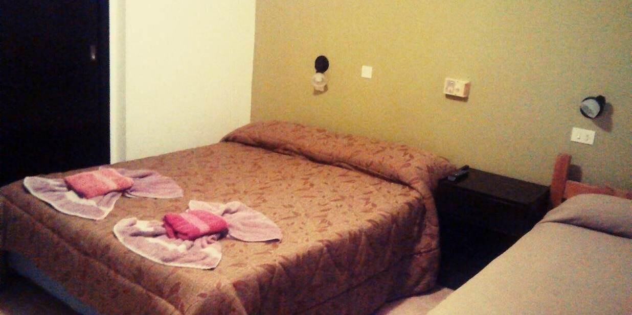 Un lit simple avec une couvre-lit à motifs et deux serviettes dessus. Il y a une table de chevet avec une lampe et une prise électrique au mur.