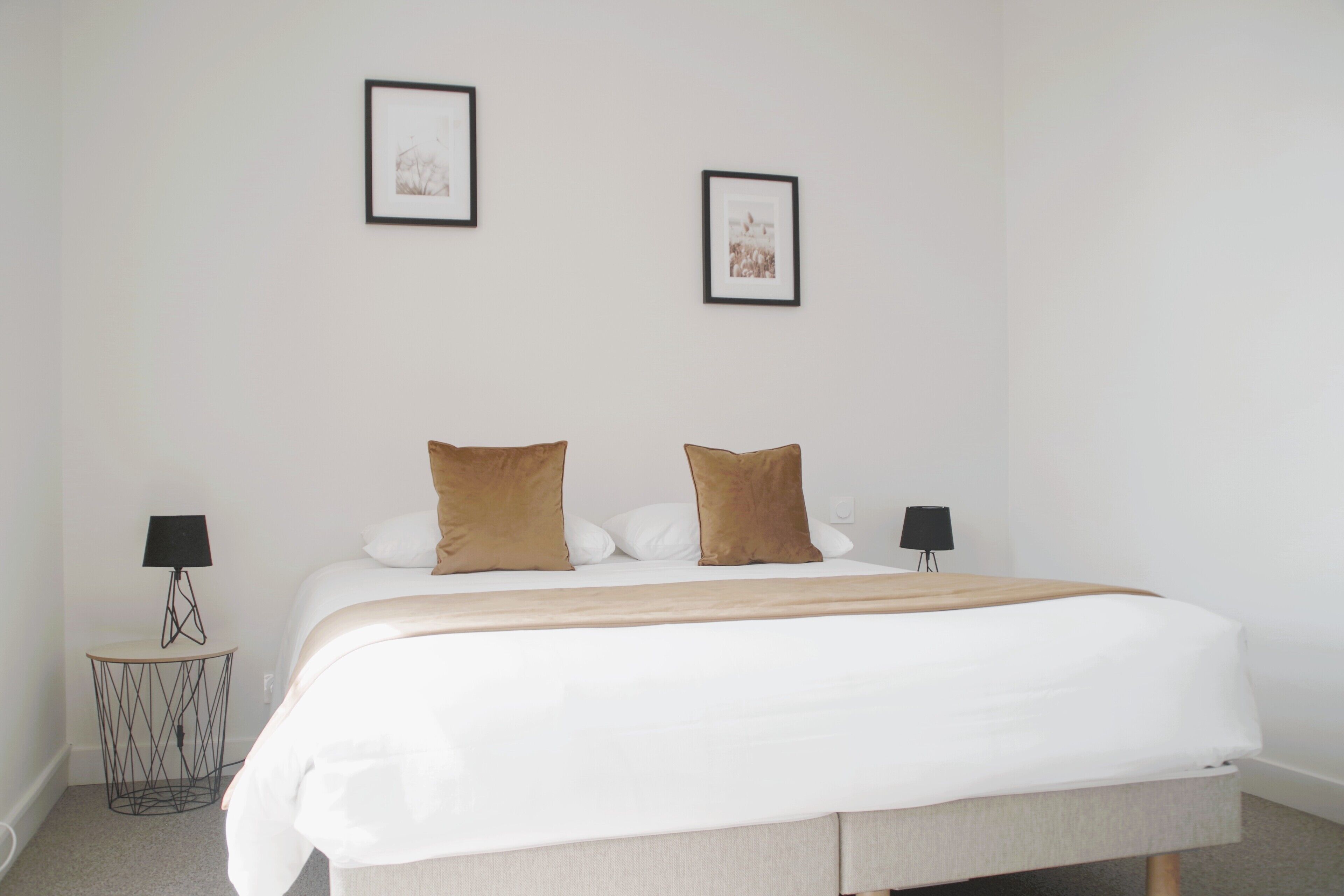 Una cama bien hecha con edredón blanco y dos almohadas marrones, una mesita de noche con lámpara y dos cuadros en la pared.