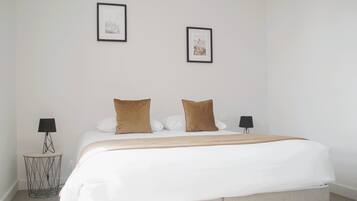 Una cama bien hecha con edredón blanco y dos almohadas marrones, una mesita de noche con lámpara y dos cuadros en la pared.