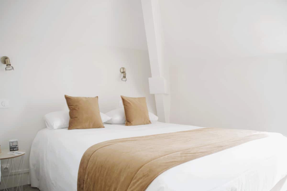 A neatly made bed with white and beige bedding, a small bedside table with a lamp, and a wall-mounted light fixture.