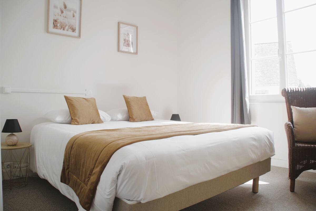 A neatly made bed with white and brown bedding, a small bedside table with a lamp, and a wicker chair.