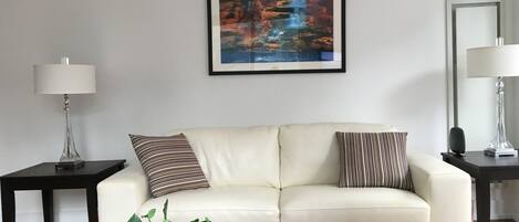 Una sala de estar moderna con un sofá blanco, dos mesitas auxiliares, un cuadro enmarcado, una planta en maceta, revistas y un control remoto sobre una mesa de centro.