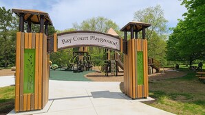 Ingang van een speeltuin, met een bord waarop 'Bay Court Playground' staat en een speeltoestel op de achtergrond.