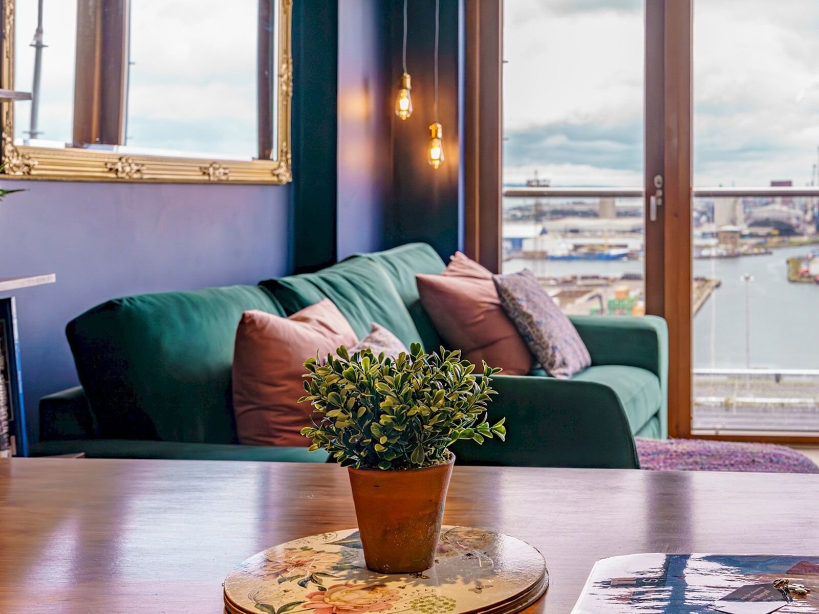 Un salon chaleureux avec un canapé vert, une plante en pot posée sur une table ronde et une vue sur une marina, visible à travers de grandes fenêtres.