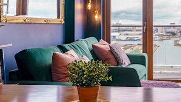 Un salon chaleureux avec un canapé vert, une plante en pot posée sur une table ronde et une vue sur une marina, visible à travers de grandes fenêtres.