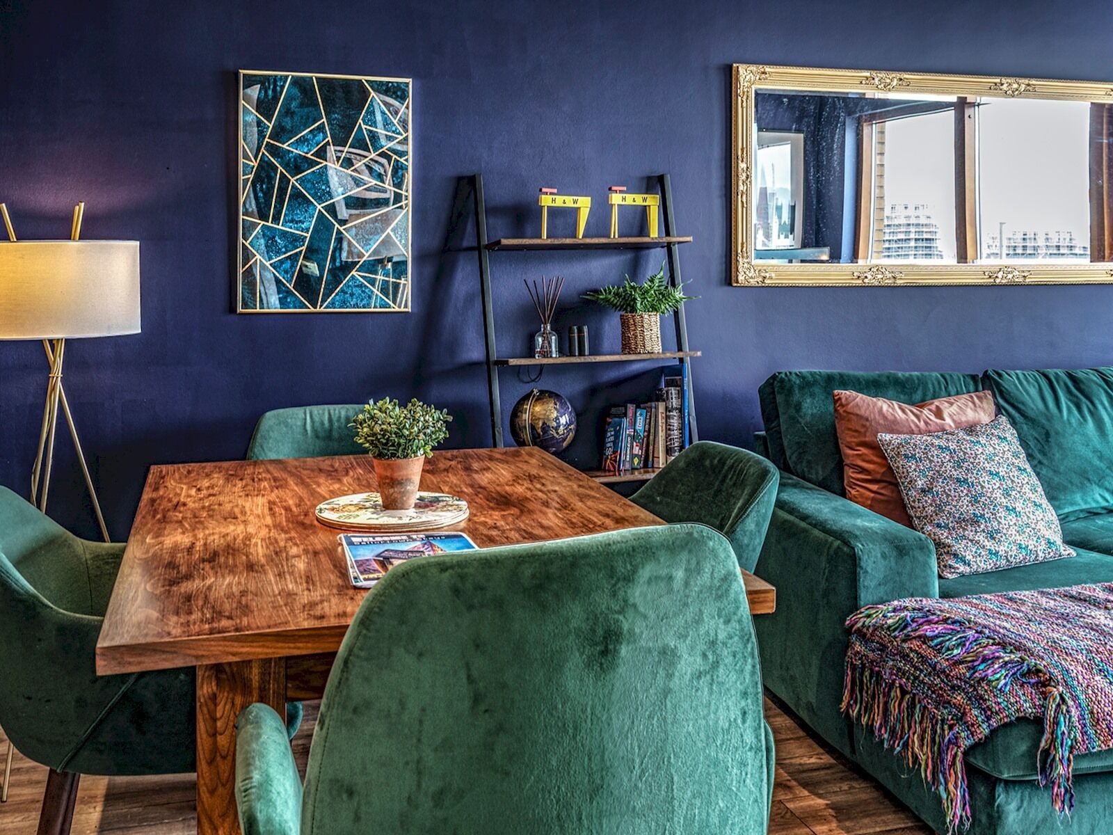 Un salon moderne avec un mur bleu foncé, une table basse en bois, des canapés en velours vert et un grand miroir qui reflète une vue sur la ville.