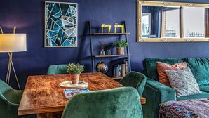 A modern living room with a dark blue wall, a wooden coffee table, green velvet sofas, and a large mirror reflecting a city view. - City Centre | Top Floor w/ Balcony & Sunset Views | Pass the Keys (Belfast)