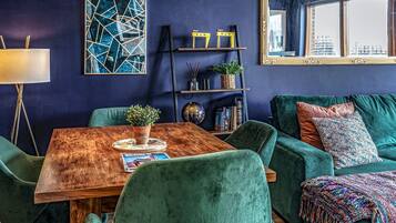 Un salon moderne avec un mur bleu foncé, une table basse en bois, des canapés en velours vert et un grand miroir qui reflète une vue sur la ville.