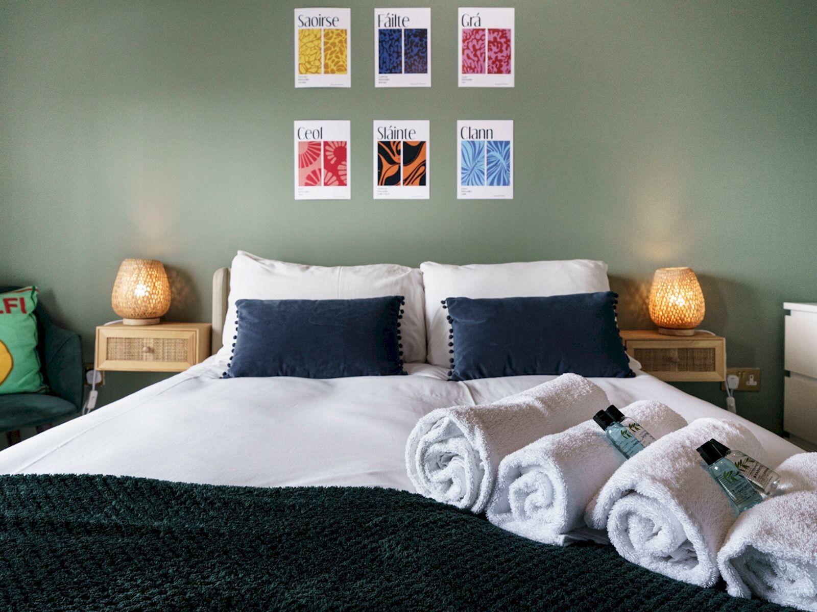 Un lit bien fait, avec des oreillers blancs et bleu marine, une couverture vert foncé et des serviettes blanches pliées, posées sur une table de chevet en bois.